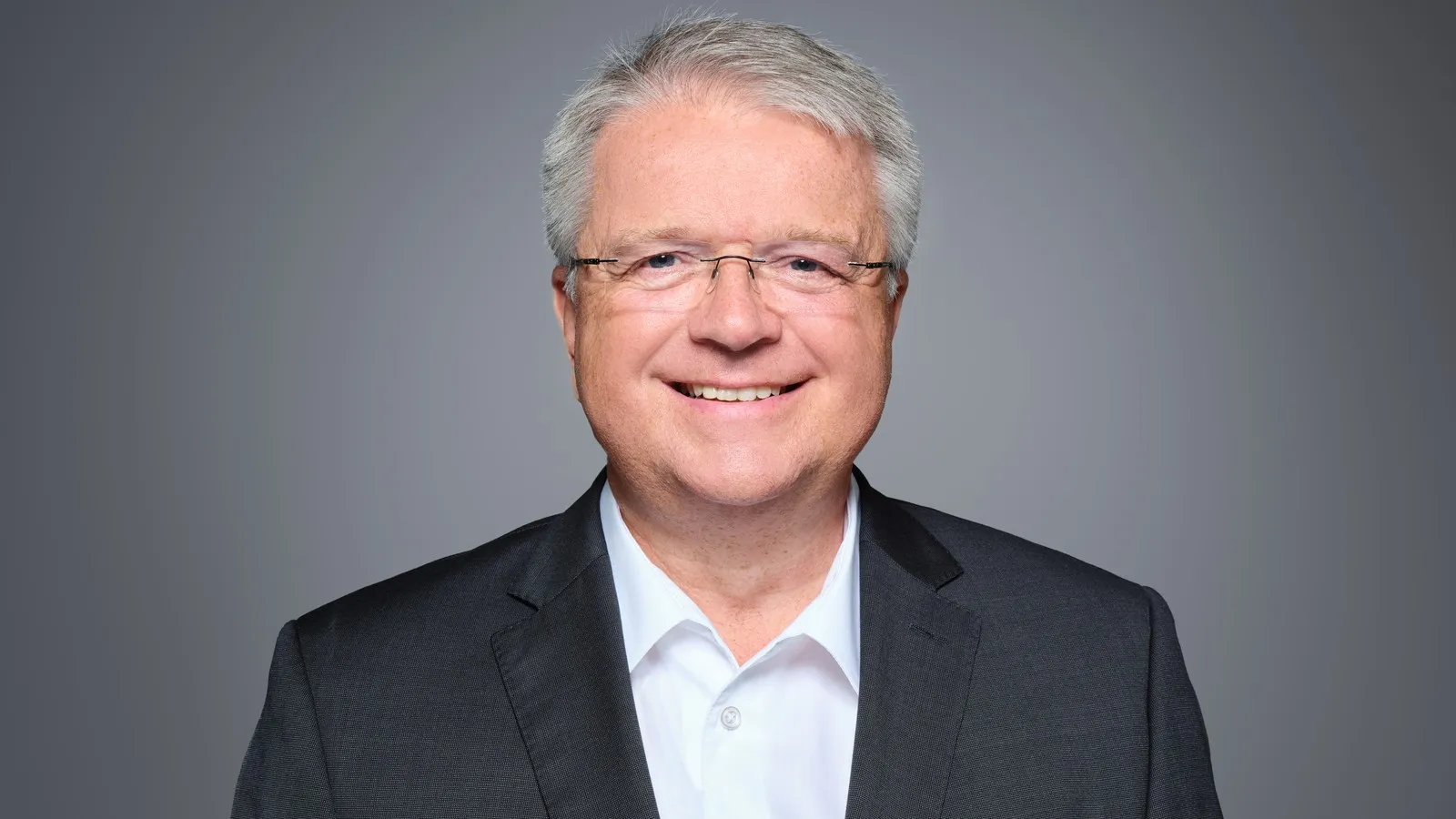 Ein weißer Mann in einem schwarzen Jackett und weißem Hemd vor grauem Hintergrund.  